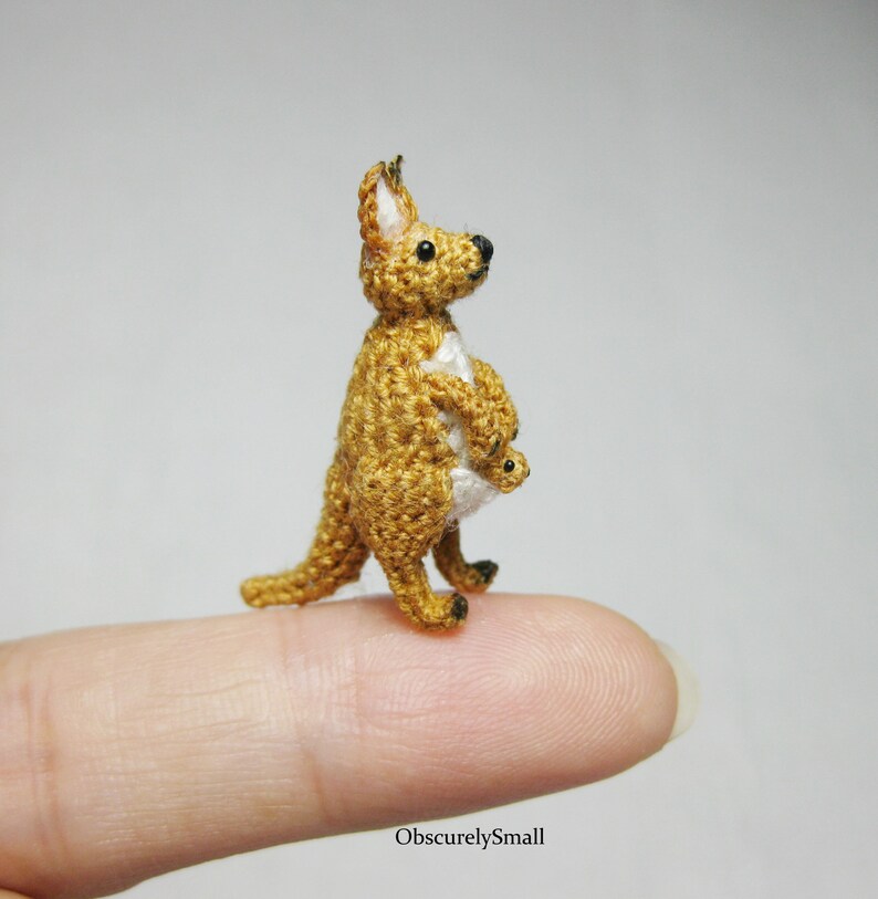 May include: A small crocheted kangaroo with a joey in its pouch. The kangaroo is brown and white, and the joey is white. The kangaroo is standing on a finger.