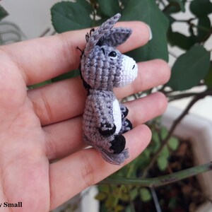 Puede incluir: Un peque&ntilde;o burro de crochet gris con pezu&ntilde;as negras y una mancha blanca en el costado. El burro mide aproximadamente 5 cm de alto y est&aacute; siendo sostenido en la mano de una persona.