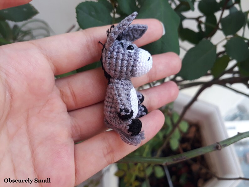 Puede incluir: Un peque&ntilde;o burro de crochet gris con pezu&ntilde;as negras y una mancha blanca en el costado. El burro mide aproximadamente 5 cm de alto y est&aacute; siendo sostenido en la mano de una persona.