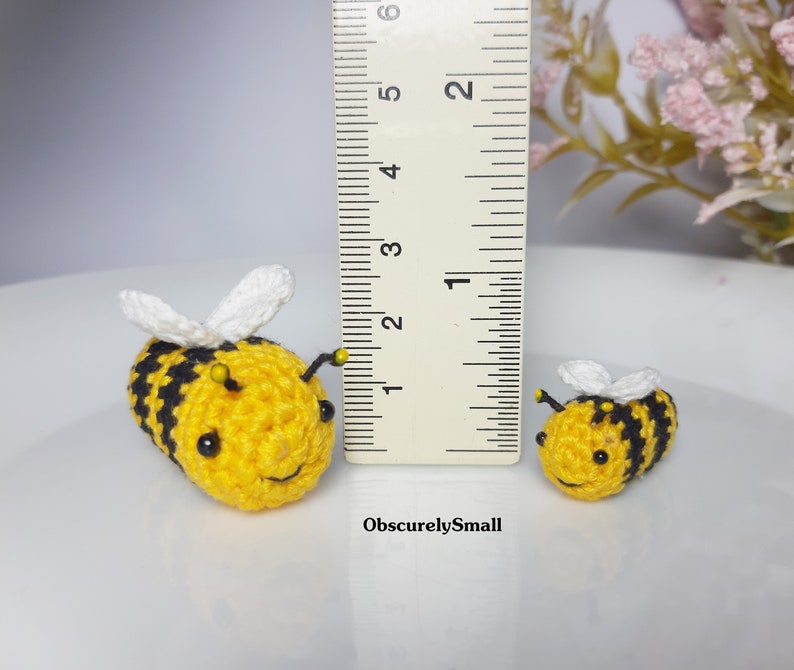 Op de afbeelding: Twee gehaakte hommels, een groter dan de andere, worden getoond naast een liniaal. De grotere hommel is geel en zwart met witte vleugels. De kleinere hommel is geel en zwart met witte vleugels. De liniaal is gemarkeerd in inches.