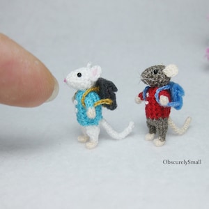 May include: Two small crocheted mice wearing backpacks. One mouse is white with a blue backpack and the other is gray with a red backpack.