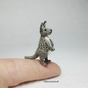 May include: A small crocheted kangaroo with a joey in its pouch. The kangaroo is gray and white and is standing on a finger.