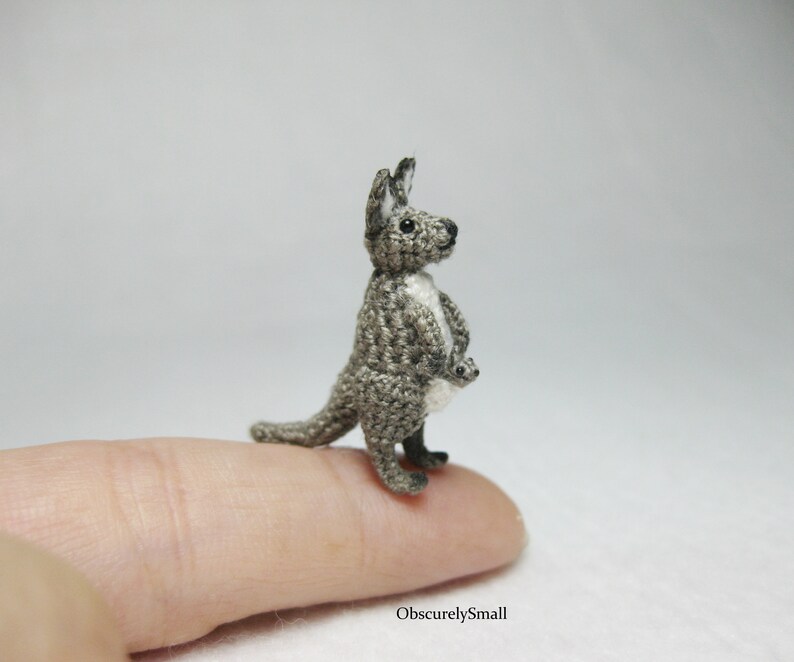 May include: A small crocheted kangaroo with a joey in its pouch. The kangaroo is gray and white and is standing on a finger.