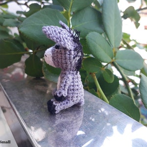 Puede incluir: Un peque&ntilde;o burro de crochet gris sentado en una superficie plateada. El burro tiene ojos negros y una crin negra.