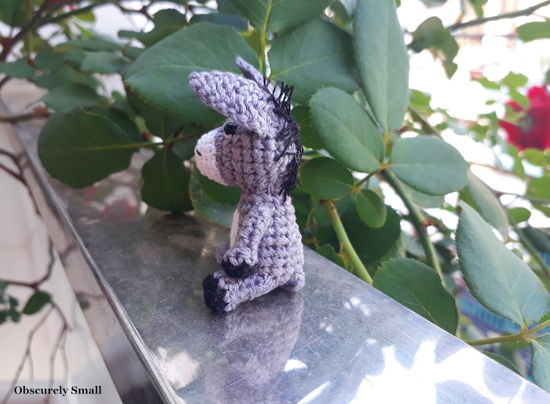 Puede incluir: Un peque&ntilde;o burro de crochet gris sentado en una superficie plateada. El burro tiene ojos negros y una crin negra.