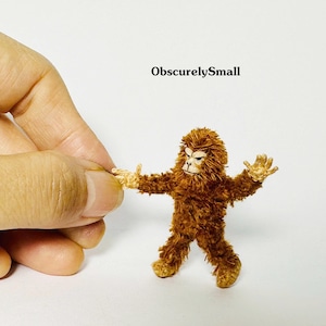 May include: A miniature brown crocheted creature with a textured, fuzzy appearance, light tan hands and feet, and a detailed face. The text ObscurelySmall is printed above the figure. The figure is shown in a standing pose with arms outstretched.