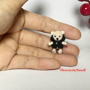 Op de afbeelding: Een miniatuur, met de hand gehaakte teddybeer met een crèmekleurig lichaam en een zwarte jas. De kleine beer heeft zwarte knoopogen en is op de hand van een persoon geplaatst. De tekst "ObscurelySmall" is zichtbaar.