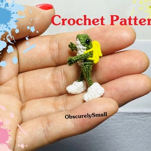 Puede incluir: Una rana verde de crochet con una mochila amarilla y zapatos blancos. La rana está siendo sostenida en la mano de una persona. La imagen tiene el texto "Crochet Pattern" y "ObscurelySmall".