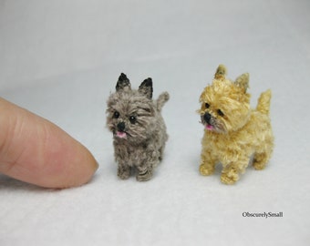 May include: Two miniature Cairn Terrier dogs made of yarn. One is gray and the other is light brown. They are both standing on a white surface.