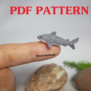 May include: A crocheted gray shark, approximately 2 inches long, is resting on a finger. The shark has a white underside and a small, pointed fin. The image includes the text "PDF PATTERN" in red letters at the top of the image.