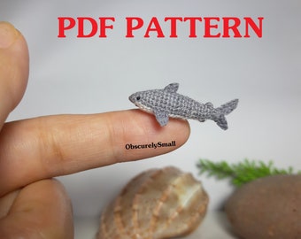 May include: A crocheted gray shark, approximately 2 inches long, is resting on a finger. The shark has a white underside and a small, pointed fin. The image includes the text "PDF PATTERN" in red letters at the top of the image.