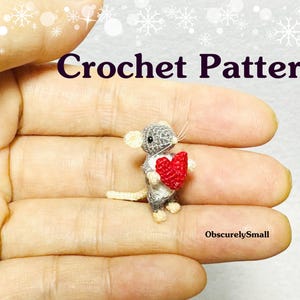May include: A crocheted grey mouse holding a red heart. The mouse is small and is being held in a person's hand. The text "Crochet Pattern" is at the top of the image and the text "ObscurelySmall" is at the bottom of the image.