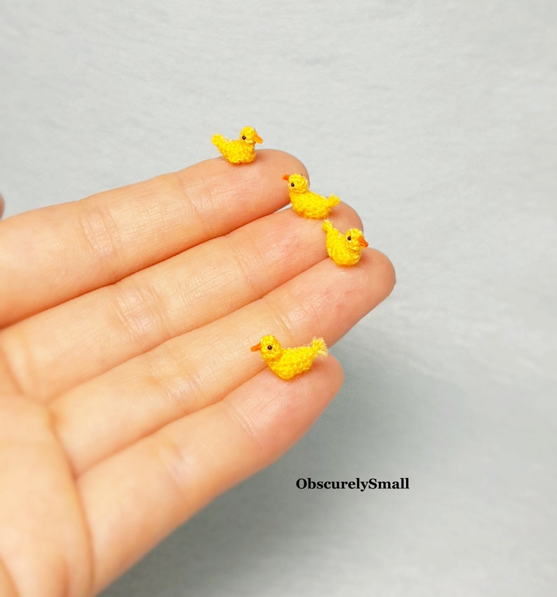 May include: Four tiny crocheted yellow ducklings with orange beaks and feet, arranged in a line on a hand.