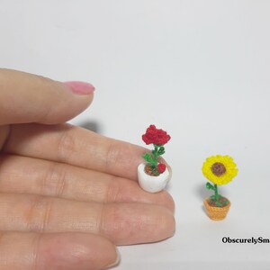 May include: Miniature crochet flowers, including a red rose in a white pot and a yellow sunflower in a brown pot. The flowers are handmade and detailed, showcasing a delicate craft. The text "ObscurelySmall" is visible.