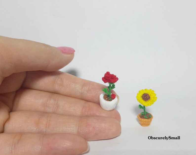 May include: Miniature crochet flowers, including a red rose in a white pot and a yellow sunflower in a brown pot. The flowers are handmade and detailed, showcasing a delicate craft. The text "ObscurelySmall" is visible.