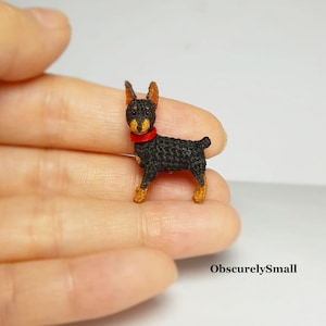 May include: A small crocheted black and brown dog with a red collar. The dog is standing on a hand.