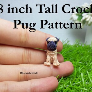Puede incluir: Un perro pug de crochet, de aproximadamente 2 cm de alto, está sostenido en la mano de una persona. El pug es beige con rasgos faciales negros y está sentado sobre una superficie de hierba verde.