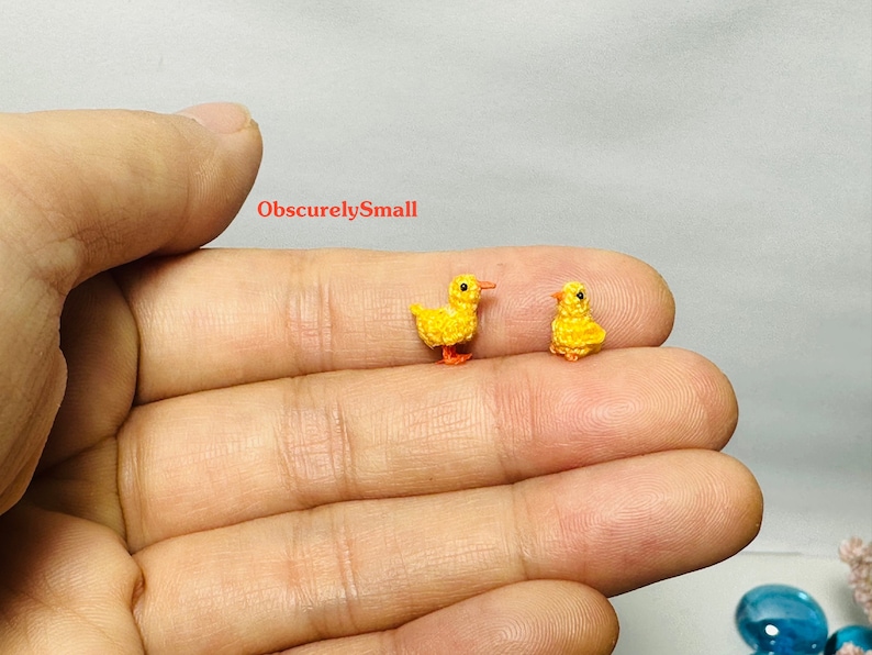 May include: Two small crocheted yellow ducklings with orange beaks and feet. The ducklings are sitting on a person's hand.