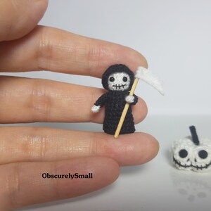 May include: A small crocheted Grim Reaper figure holding a scythe, with a white crocheted pumpkin in the background.