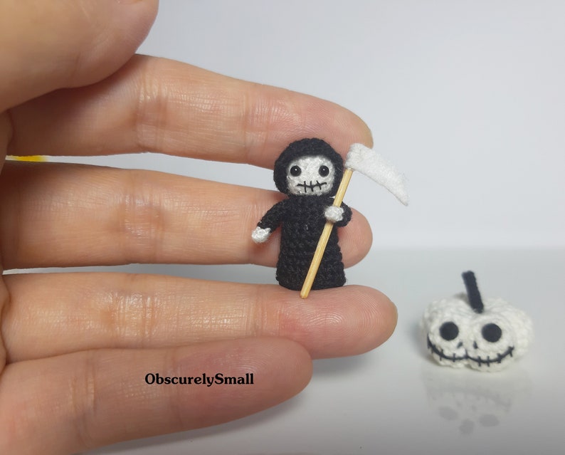 May include: A small crocheted Grim Reaper figure holding a scythe, with a white crocheted pumpkin in the background.