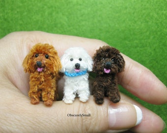 May include: Three miniature crocheted poodle dogs, one brown, one white with a blue collar, and one dark brown, sitting on a hand.