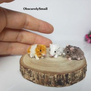 May include: Three crocheted hamster figurines, one orange and white, one white, and one gray. The figurines are sitting on a wooden slice.