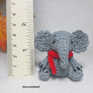 May include: A small gray crocheted elephant figurine wearing a red scarf, positioned next to a ruler showing a height of approximately 2 inches. The elephant has black bead eyes and black stitched details on its feet. The text ObscurelySmall is visible.