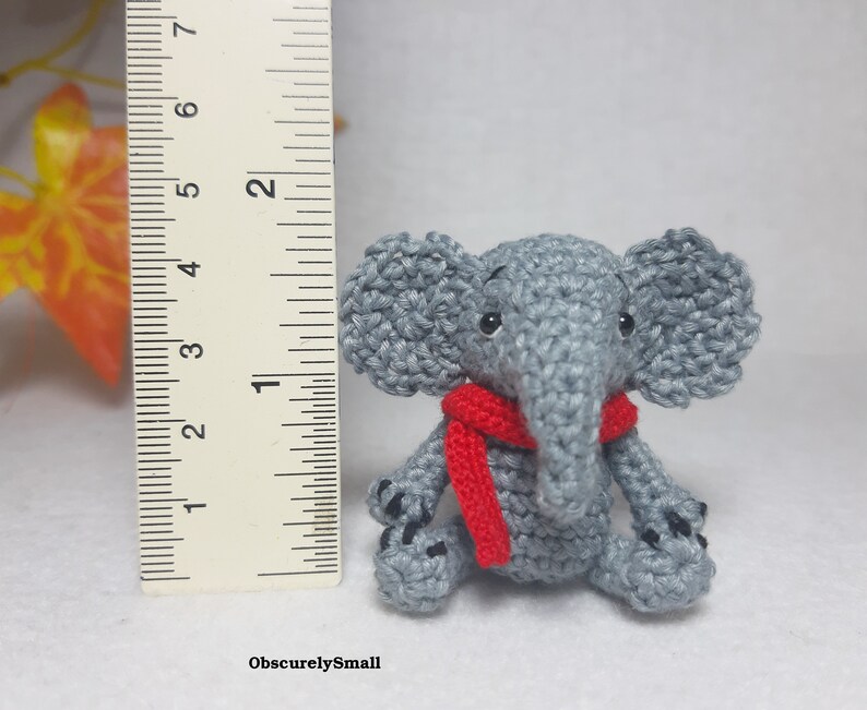 May include: A small gray crocheted elephant figurine wearing a red scarf, positioned next to a ruler showing a height of approximately 2 inches. The elephant has black bead eyes and black stitched details on its feet. The text ObscurelySmall is visible.