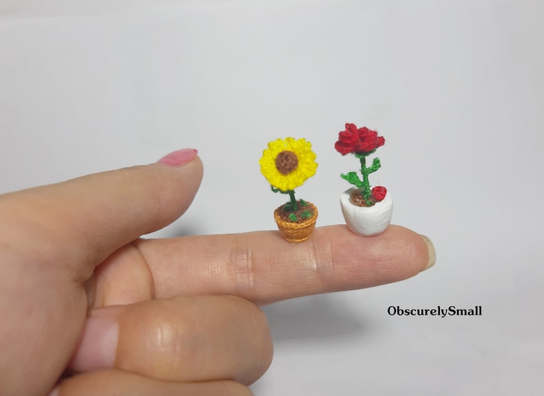 May include: Two miniature crochet potted plants. One is a yellow sunflower in a brown pot, the other is a red rose in a white pot. The plants are displayed on a finger. The text "ObscurelySmall" is in the bottom right corner.