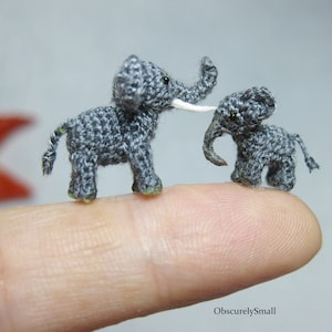 May include: Two tiny crocheted gray elephants, one larger than the other, standing on a finger. The larger elephant has a white tusk.
