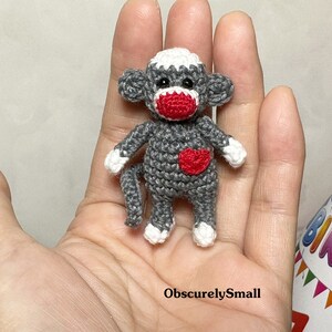 Puede incluir: Un peque&ntilde;o mono de crochet con pelaje gris, cara blanca y nariz y mejillas rojas. El mono lleva un coraz&oacute;n rojo en el pecho.