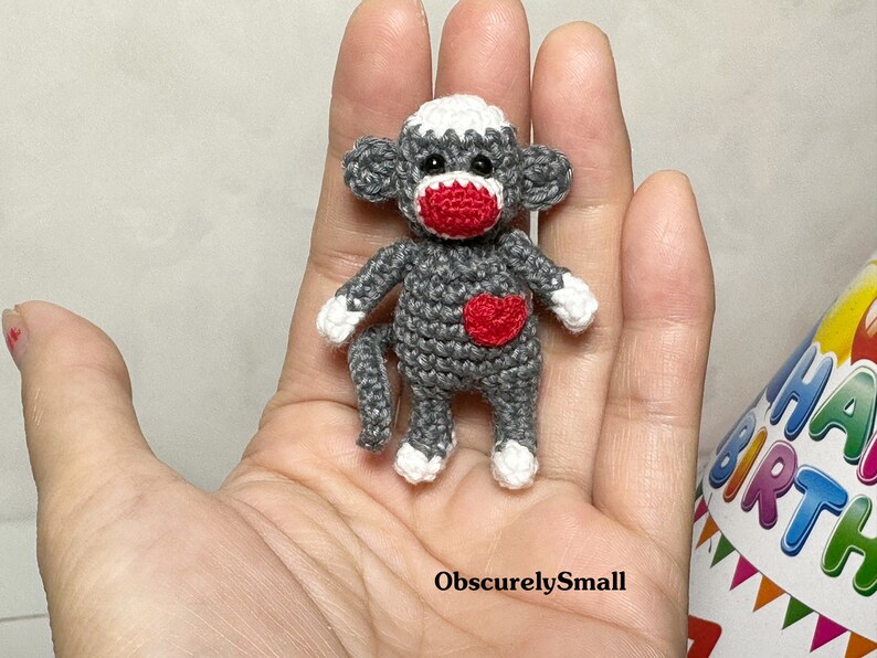 Puede incluir: Un peque&ntilde;o mono de crochet con pelaje gris, cara blanca y nariz y mejillas rojas. El mono lleva un coraz&oacute;n rojo en el pecho.