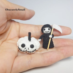 May include: A hand holding two crocheted Halloween decorations. One is a white skull with black eyes and a black mouth. The other is a black Grim Reaper with a white scythe and a wooden handle.