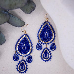 Könnte beinhalten: Ein Paar handgefertigte blaue Perlenohrringe mit goldfarbenen Akzenten. Die Ohrringe haben ein tropfenförmiges Design mit kleineren, herabhängenden Tropfen. Die aufwendige Perlenarbeit und die goldenen Haken verleihen dem Schmuck Eleganz.