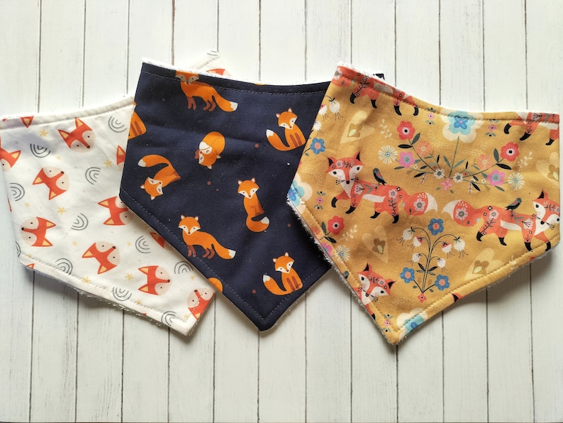 May include: Three fox-themed baby bibs. One bib is white with a light blue and white pattern, one is navy blue with a white pattern, and one is yellow with a floral pattern.