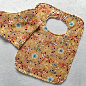 May include: A yellow bib with a fox and floral pattern. The bib has a snap closure and a matching burp cloth.