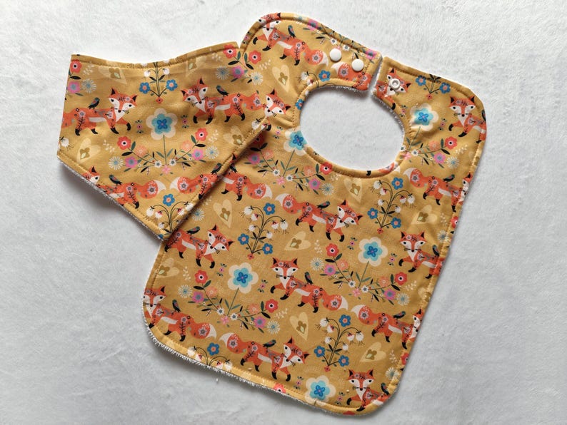 May include: A yellow bib with a fox and floral pattern. The bib has a snap closure and a matching burp cloth.