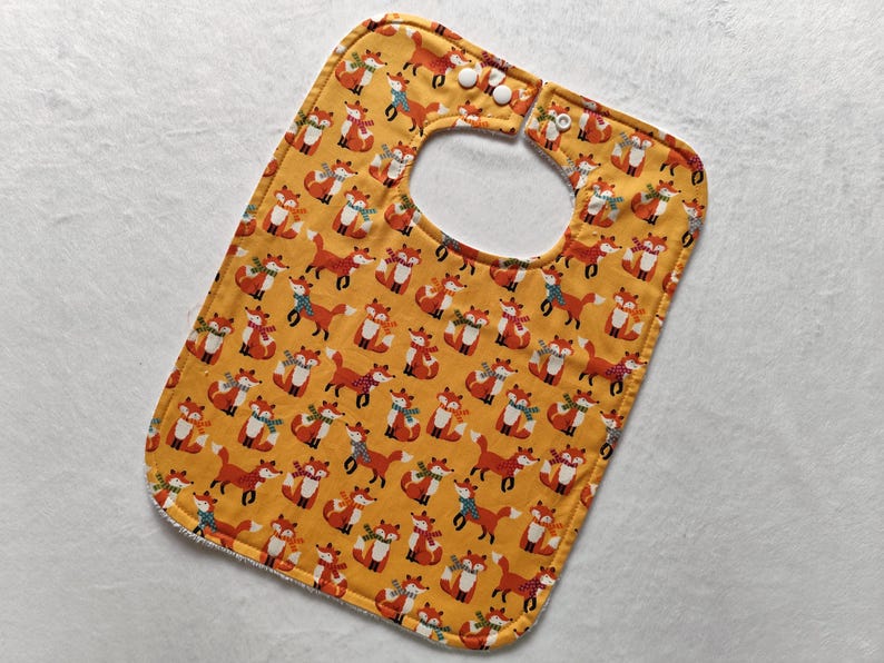 May include: A yellow baby bib with a fox print. The bib has two white snaps on the neck.