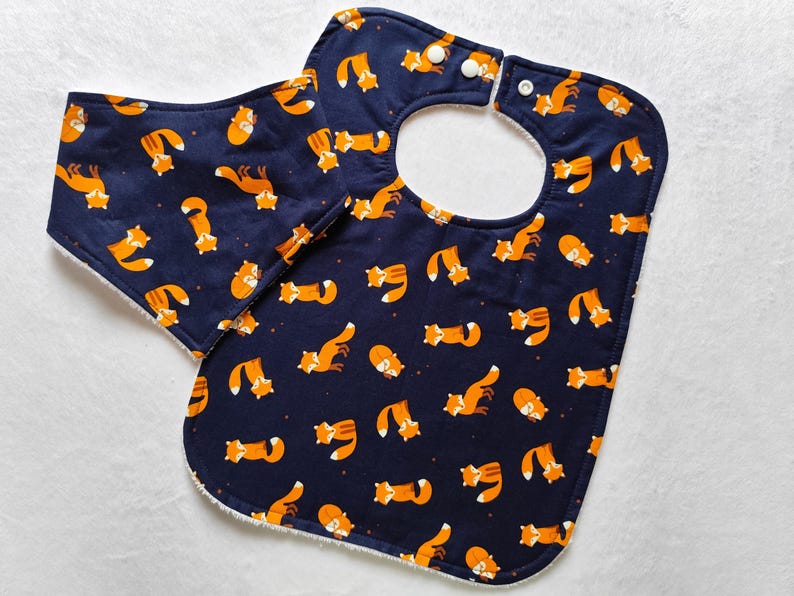 May include: A blue and white baby bib with a fox print. The bib has a white fleece lining and a snap closure.