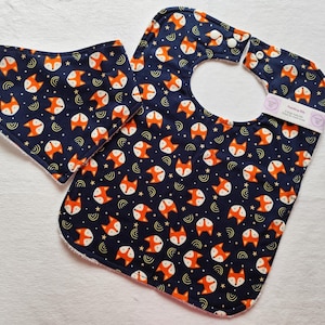 May include: A blue and orange bib with a fox print and a matching smaller bib. The bib has a white snap closure.