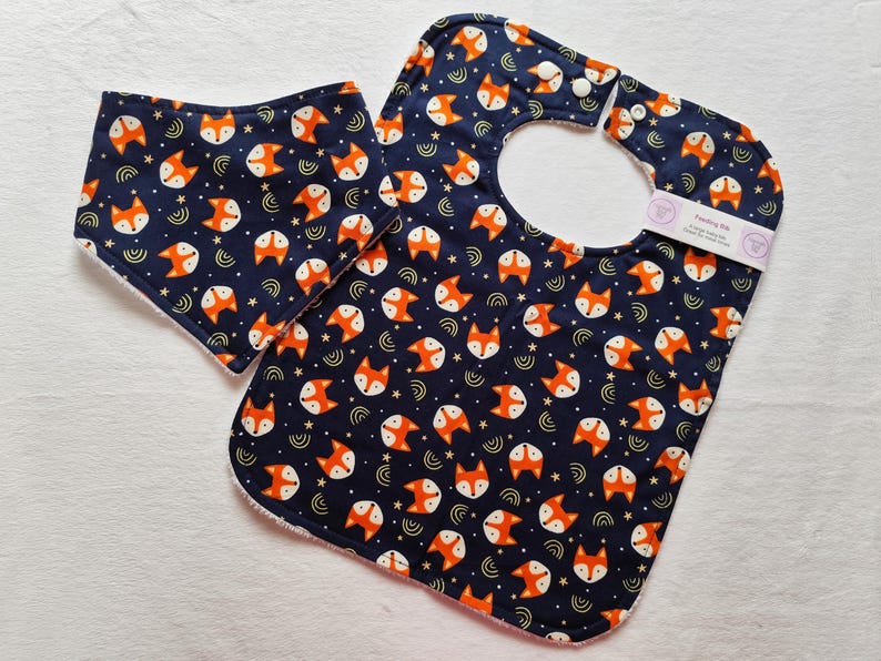 May include: A blue and orange bib with a fox print and a matching smaller bib. The bib has a white snap closure.