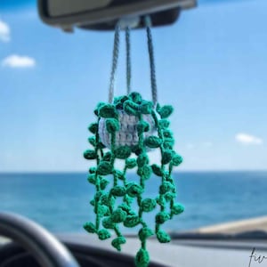 Könnte beinhalten: Eine gehäkelte Hängepflanze mit grünen Blättern und einem weißen Topf, die am Rückspiegel eines Autos aufgehängt ist. Die Pflanze befindet sich vor dem Hintergrund von Meer und Himmel. Der Text "five crochet" ist sichtbar.