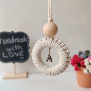 Op de afbeelding: Een witte macramé ring met een kleine Eiffeltoren bedel eraan hangend. De ring is bevestigd aan een houten kraal met een bladeren ontwerp. De ring hangt aan een wit koord.