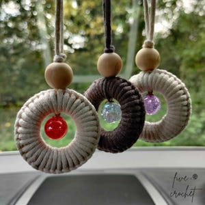 Könnte beinhalten: Drei gehäkelte Auto-Lufterfrischer mit Holzperlen und Glaskugeln. Die Kugeln sind rot, lila und schillernd. Die Lufterfrischer hängen an einem Fenster.