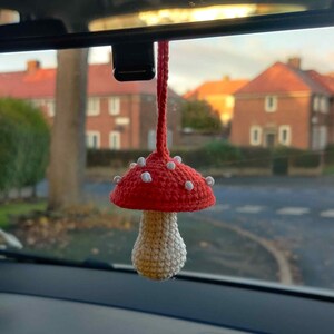 May include: A handmade crochet mushroom car charm. The mushroom has a red cap with white spots and a cream-colored stem. It hangs from a red cord, adding a whimsical touch to the car's interior.