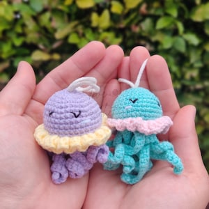 May include: Two crocheted jellyfish toys, one purple with a yellow ruffle and one teal with a pink ruffle. Both have smiling faces and are held in a person's hands.