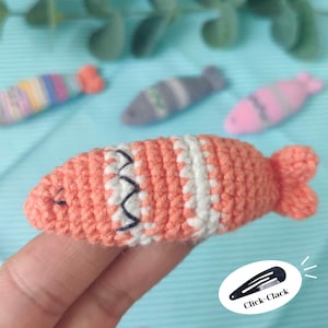 May include: A close-up of a coral-colored crochet fish hair clip with white and black accents. The clip has a fish tail and is held by a hand. The background features other fish-shaped hair clips in various colors. The text "Click-Clack" is on the bottom right.