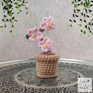 May include: A crocheted pink flower plant in a brown pot. The plant is sitting on a silver tray with an intricate design.
