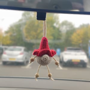 May include: A crocheted gnome car charm with a red hat and white body. The gnome is hanging from the rearview mirror of a car.