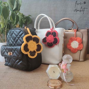 May include: Three different bags with crocheted flower charms attached to their handles. The bags are black, white, and brown. The flowers are yellow and brown, red and black, and orange and white.